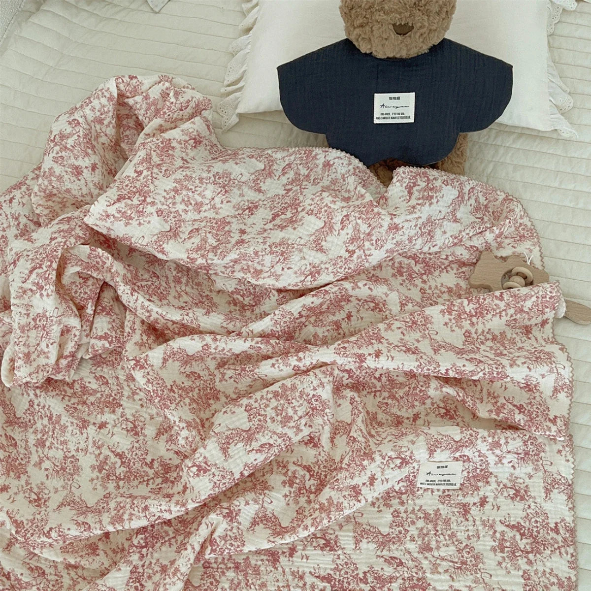 A pink floral print muslin swaddle blanket is draped next to a brown teddy bear.