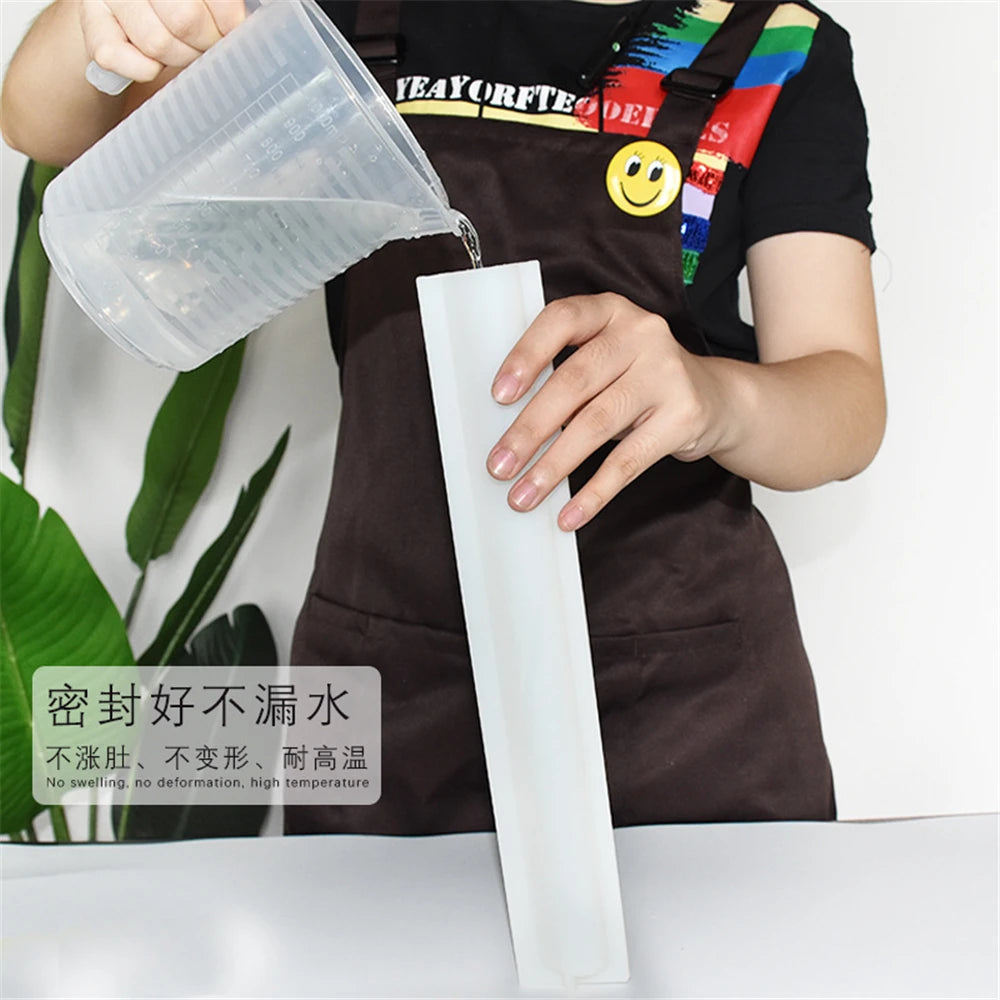 A hand pours a clear substance from a plastic mold into a white silicone mold for soap or candle making.