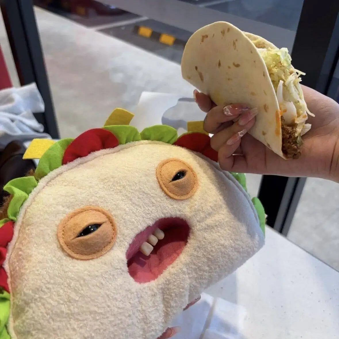 A hand holds a half-eaten soft taco in front of a cute monster plush toy with stitched features.