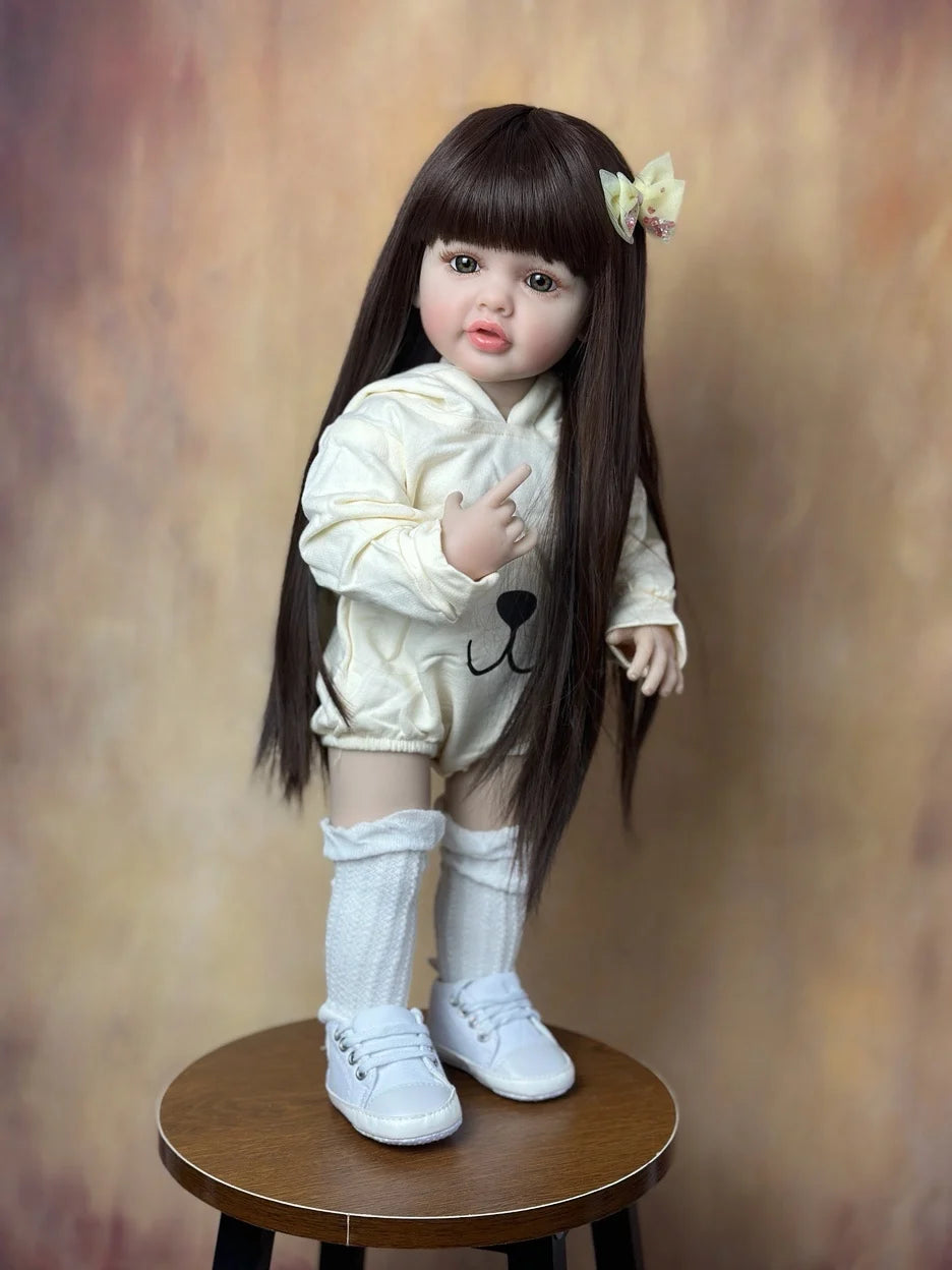 A lifelike 55 cm soft silicone baby doll with long dark hair and a white outfit standing on a wooden surface.