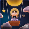 A collectible figurine of a crying girl with a yellow hat suspended by strings under a large moon in a dark night sky.