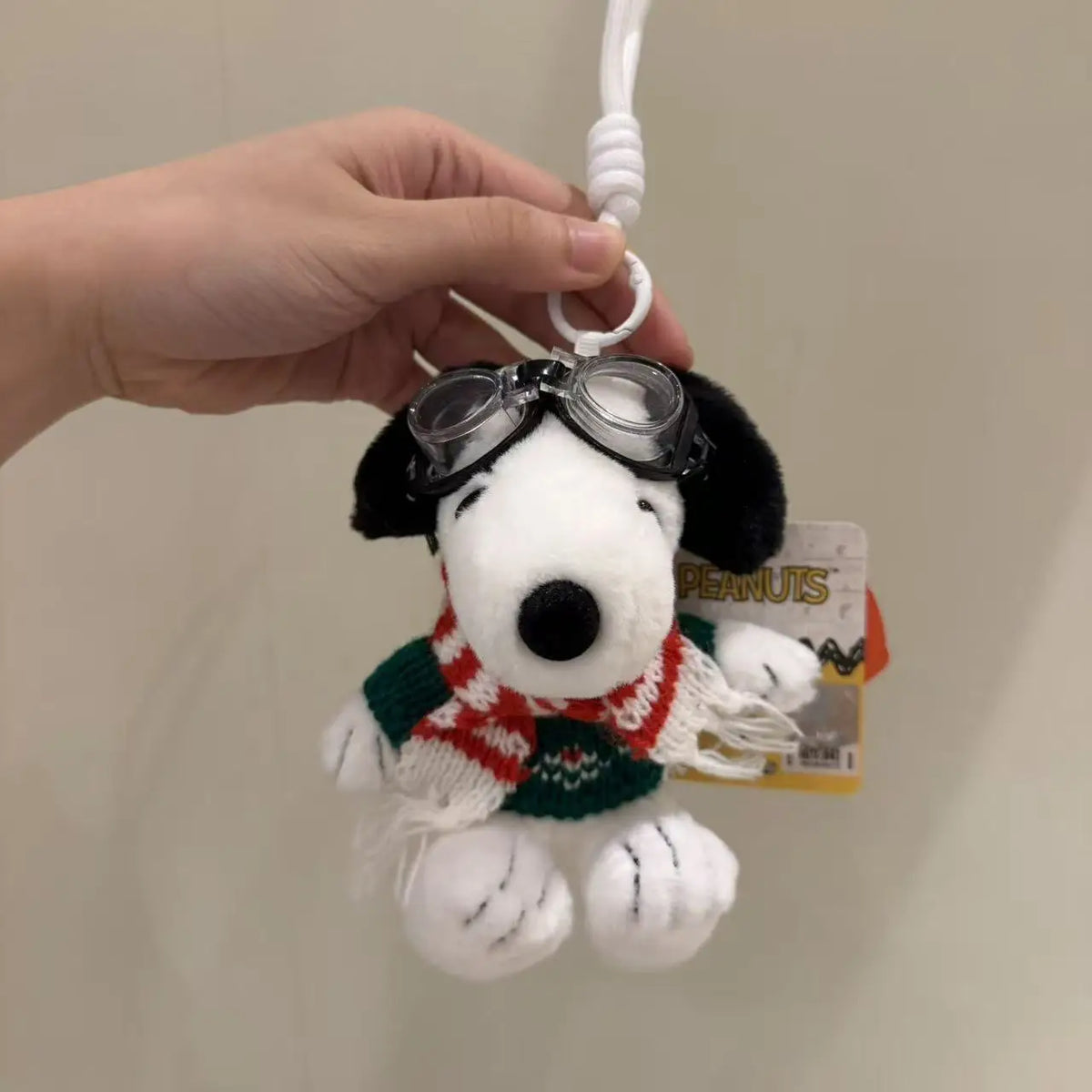 A white Snoopy plush toy wearing a red and green patterned scarf and black sunglasses is being attached to a white string.