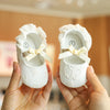 A pair of white lace-trimmed baby girl moccasin shoes with bows being held by a person.