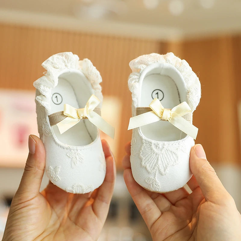 A pair of white lace-trimmed baby girl moccasin shoes with bows being held by a person.