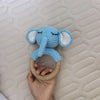 A light blue elephant rattle and teether ring made of wood is held in a hand against a quilted background.