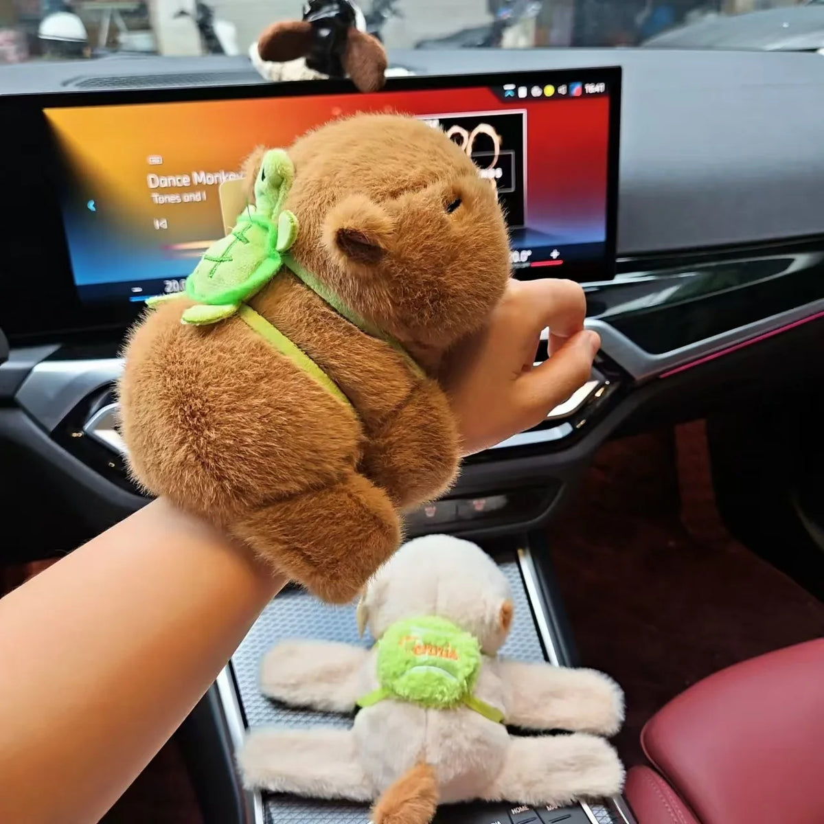 A hand holds a brown capybara plush toy wearing a green turtle friend bracelet in a car interior.