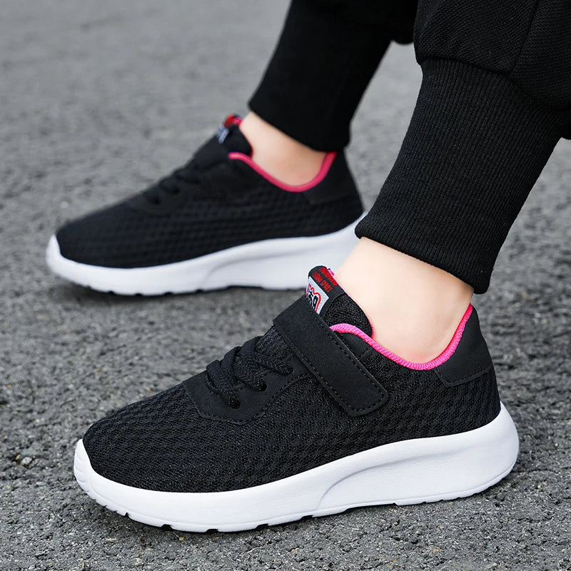 A close-up of a black breathable mesh kids sneaker with pink accents and white sole, worn on a person's foot.