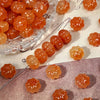 A collection of small, translucent orange resin beads shaped like pumpkins, some gold-traced, displayed on a surface.