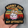Black embroidered patch with text Burned Out But Optimistic featuring a crown and sun background