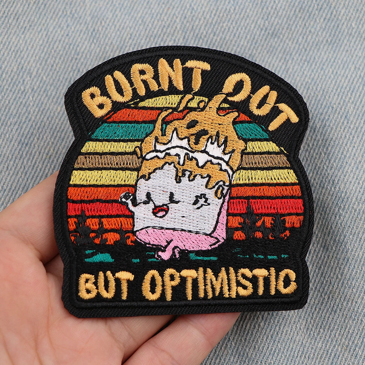 Black embroidered patch with text Burned Out But Optimistic featuring a crown and sun background