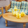 A yellow porcelain teapot and cups are displayed on a light wood table next to dollhouse furniture.
