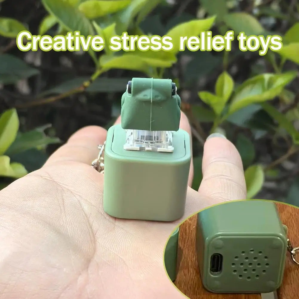 A hand holds a green LED frog croaking keychain fidget toy against a blurred green outdoor background.