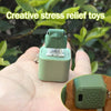 A hand holds a green LED frog croaking keychain fidget toy against a blurred green outdoor background.