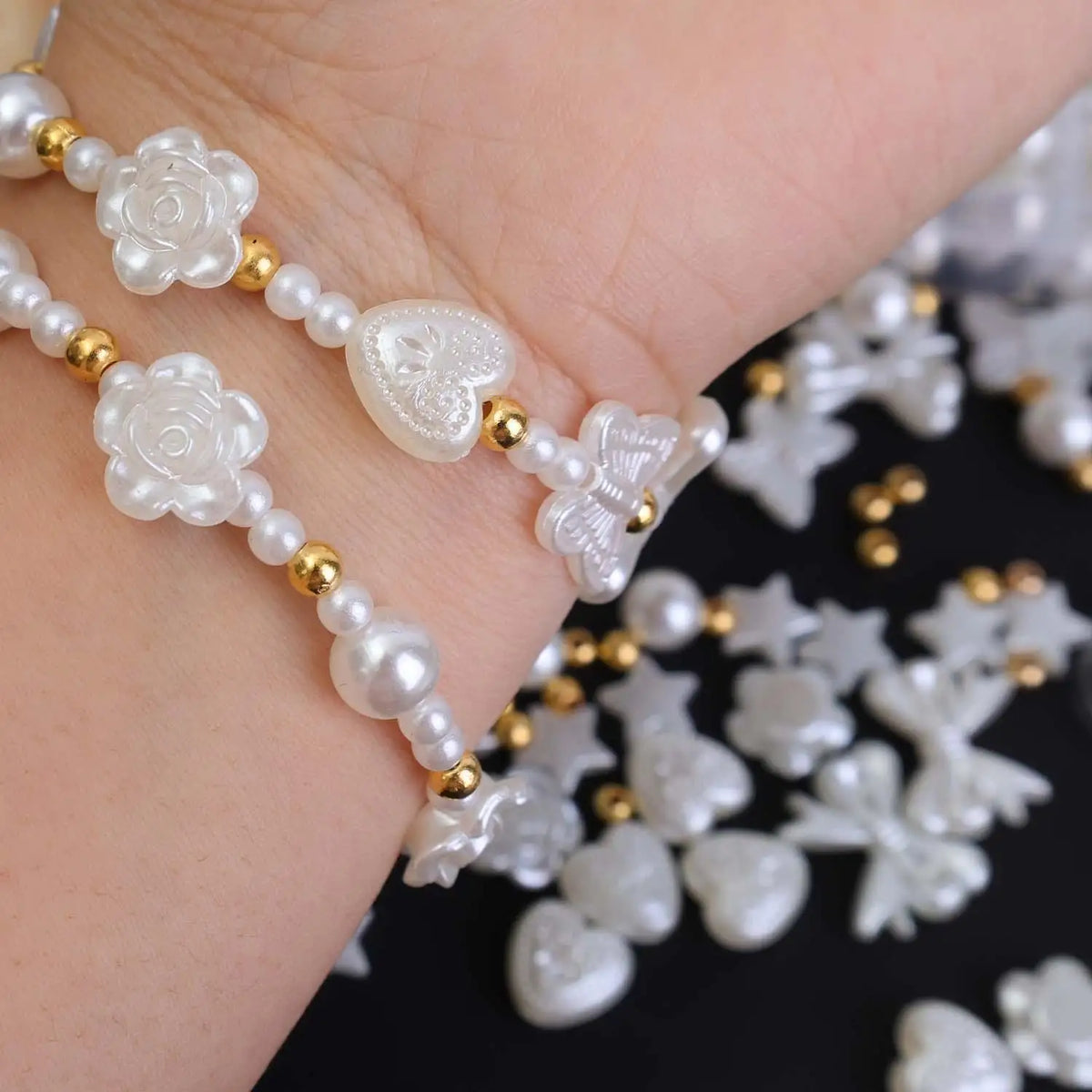 A hand is shown holding pearl bead jewelry featuring white floral and heart-shaped beads with gold accents.
