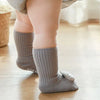A close-up of a baby's foot wearing gray non-slip cotton socks with a small animal charm on a wooden floor.
