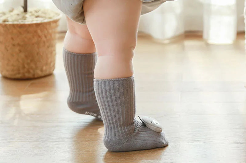 A close-up of a baby's foot wearing gray non-slip cotton socks with a small animal charm on a wooden floor.
