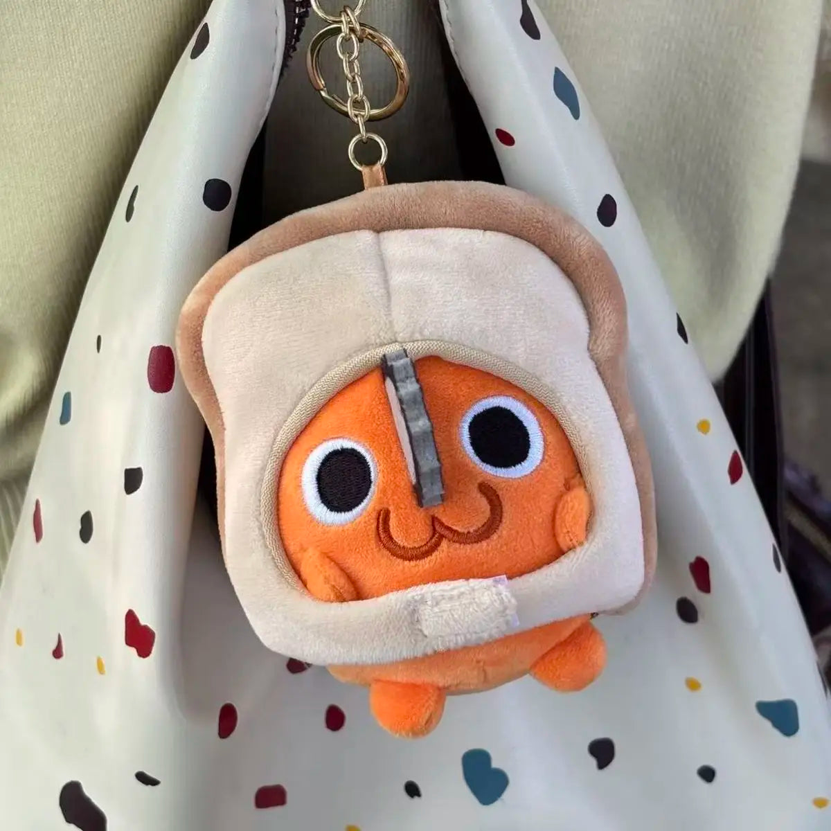 An orange Pochita plush keychain with a soft stuffed doll face is hanging from a gold chain on a light-colored fabric bag.
