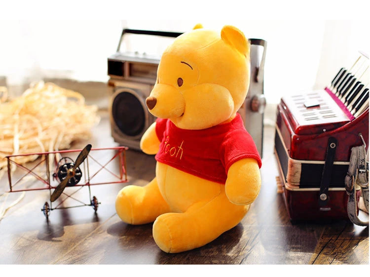 A yellow Winnie the Pooh plush bear wearing a red shirt sitting on a wooden surface next to an old radio.