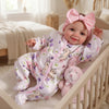 A baby doll wearing a pink bow and floral pajamas is lying in a wooden crib.