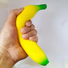 A hand is holding a yellow squishy banana stress relief toy with green accents.