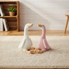 A pair of soft, stuffed goose toys in white and pink are lying together on a beige rug.