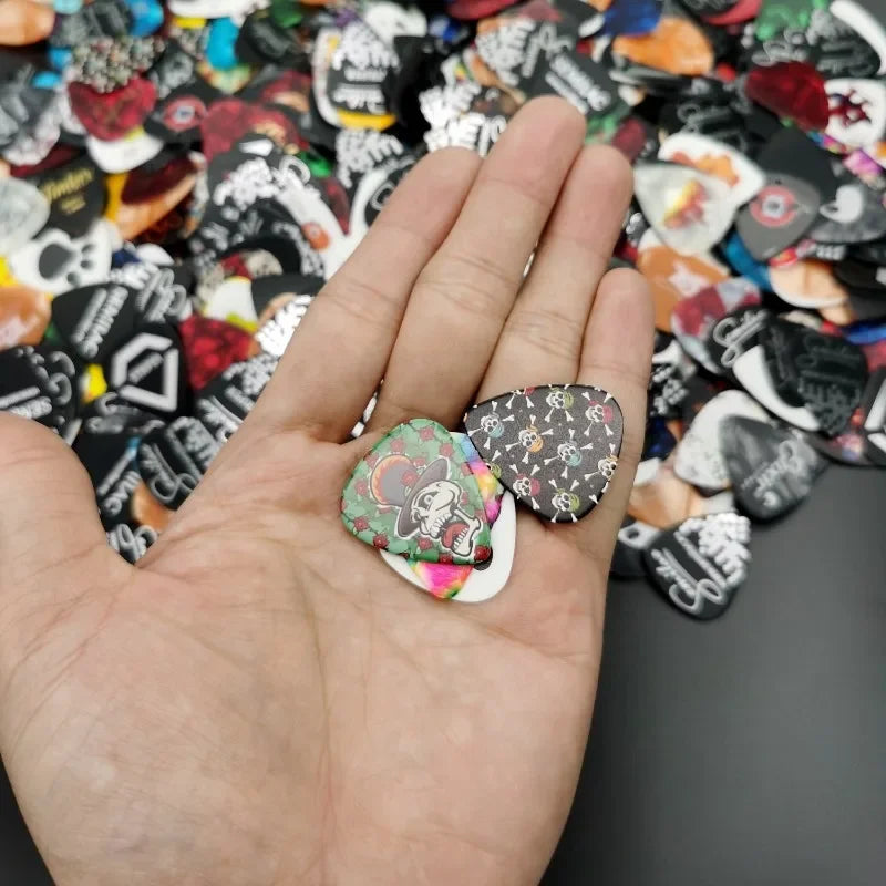 A hand holds guitar picks, some decorated with skulls and other designs, against a dark background.