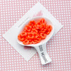 A white spoon holds bright orange, ruffled acrylic beads on a pink patterned card.