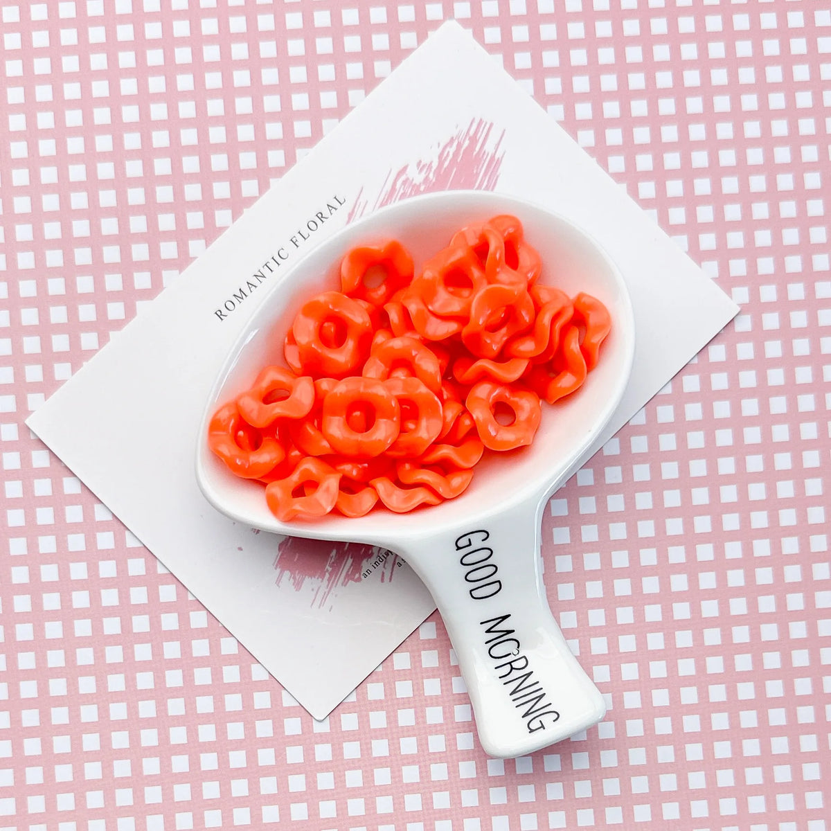 A white spoon holds bright orange, ruffled acrylic beads on a pink patterned card.