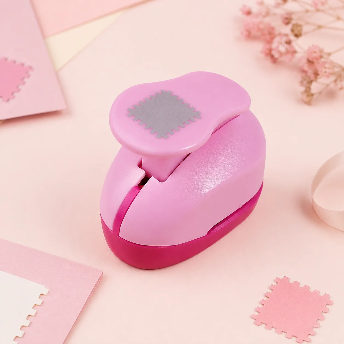 A pink decorative square stamp edge paper punch is shown on a light pink surface with other paper items.