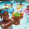 A smiling girl holds an ant-shaped water blaster toy with orange legs and a green body in a swimming pool.