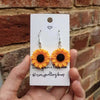 A pair of bright orange sunflower charms with black centers are displayed on a white card against a brick background.