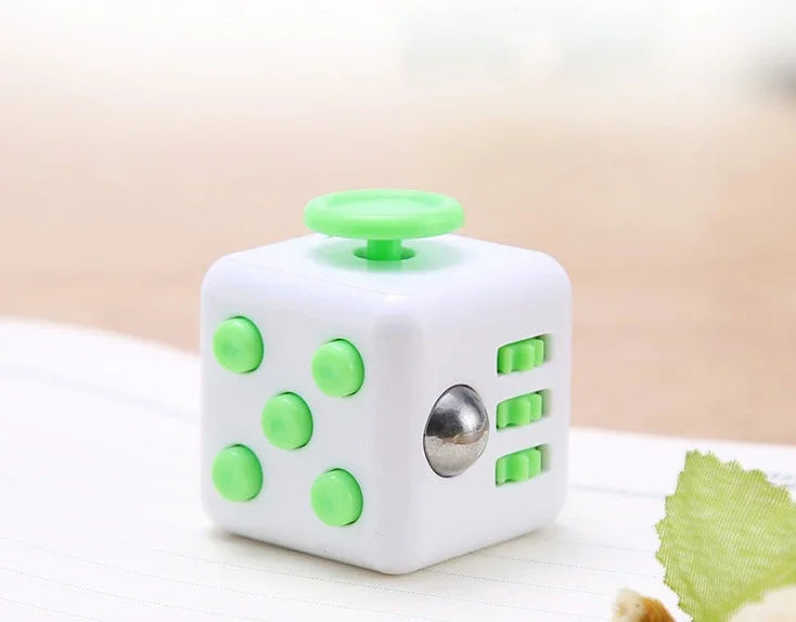 A white fidget cube with green buttons and a rotating joystick is shown on a light wooden surface.