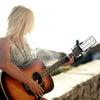 A blonde woman plays an acoustic guitar while using a 360° rotating guitar phone mount for live streaming.