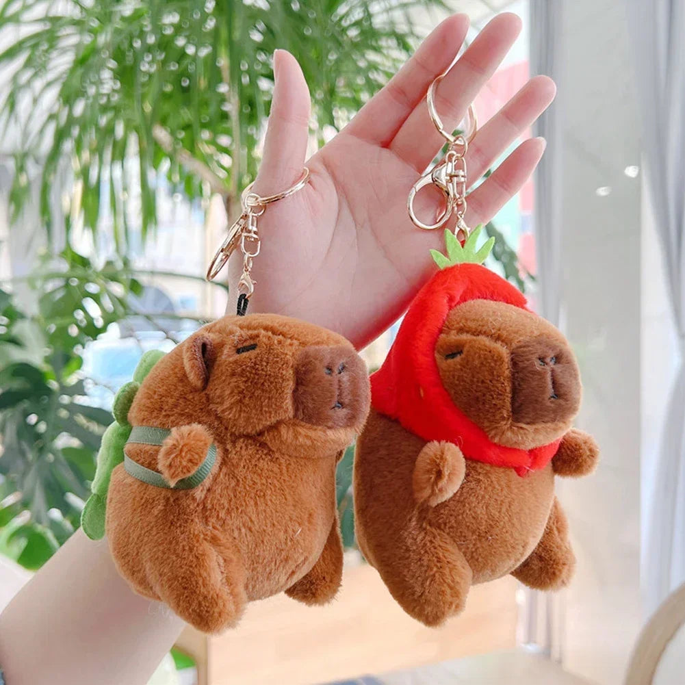 A hand holds a brown capybara plush keychain charm with a red scarf and green leaves.