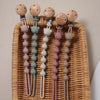 A set of silicone bear paw pacifier clips in various pastel colors hanging from a wicker tray.