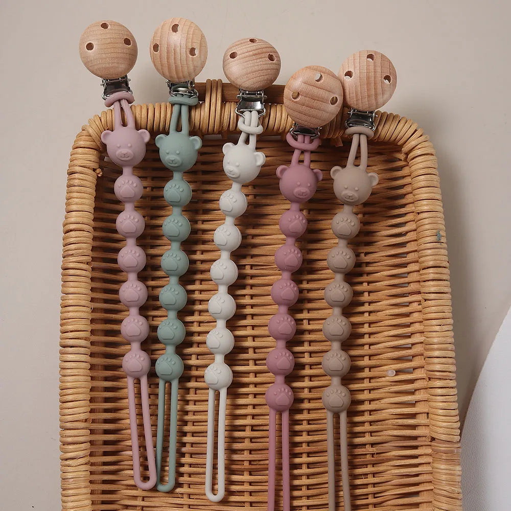 A set of silicone bear paw pacifier clips in various pastel colors hanging from a wicker tray.