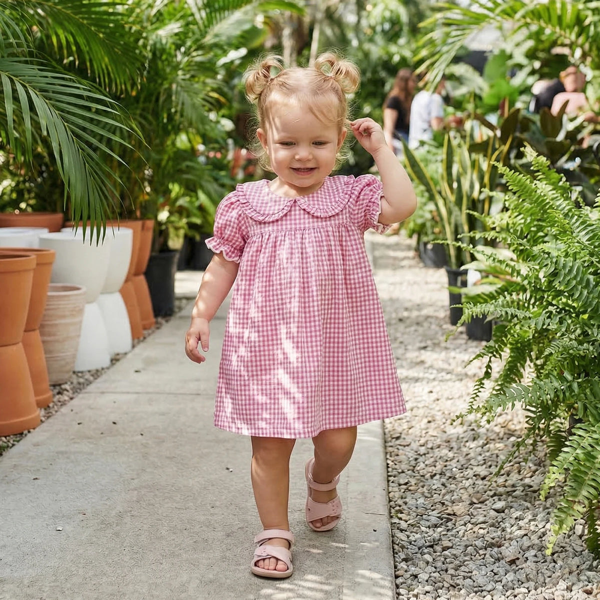 A toddler girl wearing a pink and white plaid A-line dress with a Peter Pan collar walking on a gravel path surrounded by gre