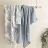 A light blue muslin swaddle blanket with a floral print is hanging on a wooden rack.