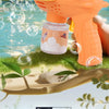 An orange bear-shaped automatic bubble gun is shown spraying bubbles near green grass and water.