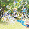A hand holds a blue bubble wand creating numerous iridescent soap bubbles in an outdoor park setting.