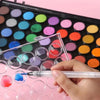 A hand dips a brush into a watercolor paint set, applying blue paint to a well in the palette.
