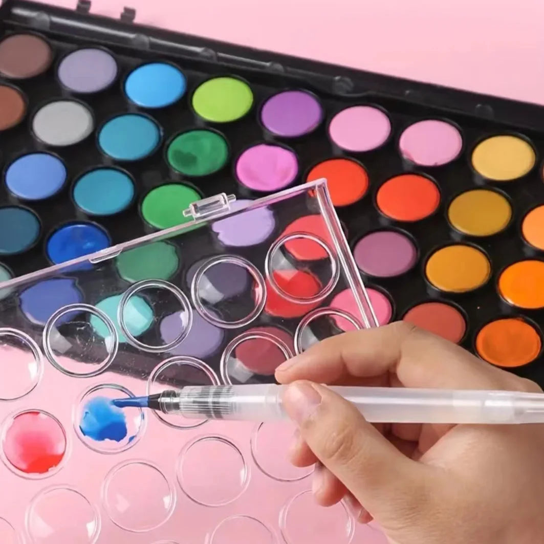A hand dips a brush into a watercolor paint set, applying blue paint to a well in the palette.