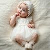 A newborn baby girl wearing a white lace tulle dress and matching hat, lying on a soft, light-colored fabric.