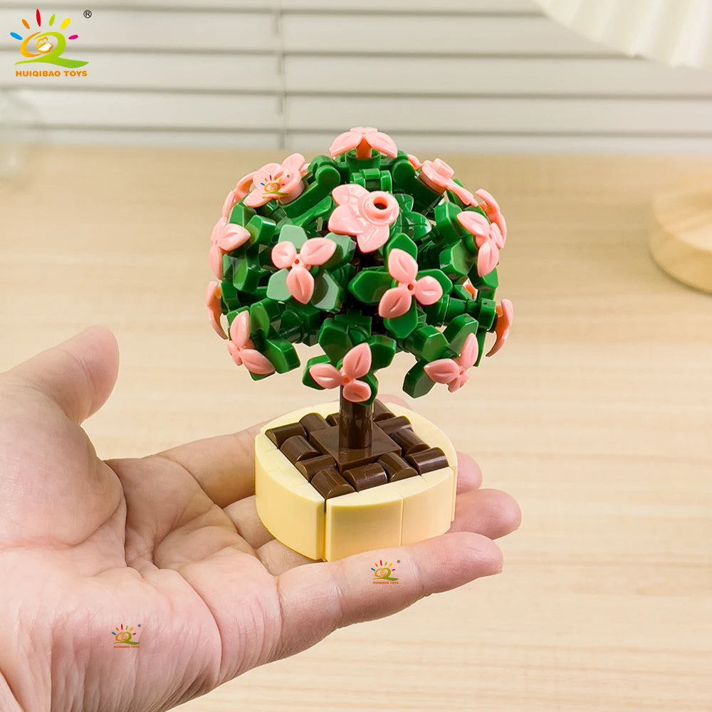 A hand holds a DIY flower succulent building blocks set shaped like a green tree with pink flowers and a brown base.