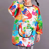 A child is wearing a colorful, long-sleeve art smock featuring handprints and paint splatters.