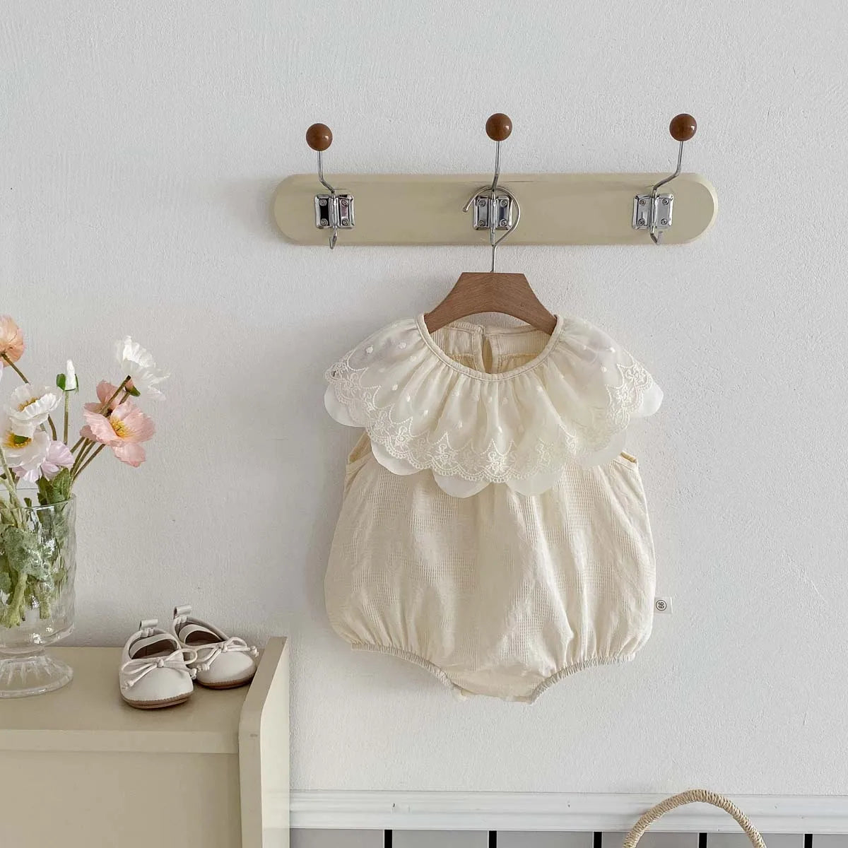 A cream-colored baby girl sleeveless jumpsuit with a lace collar hanging on a wall rack.