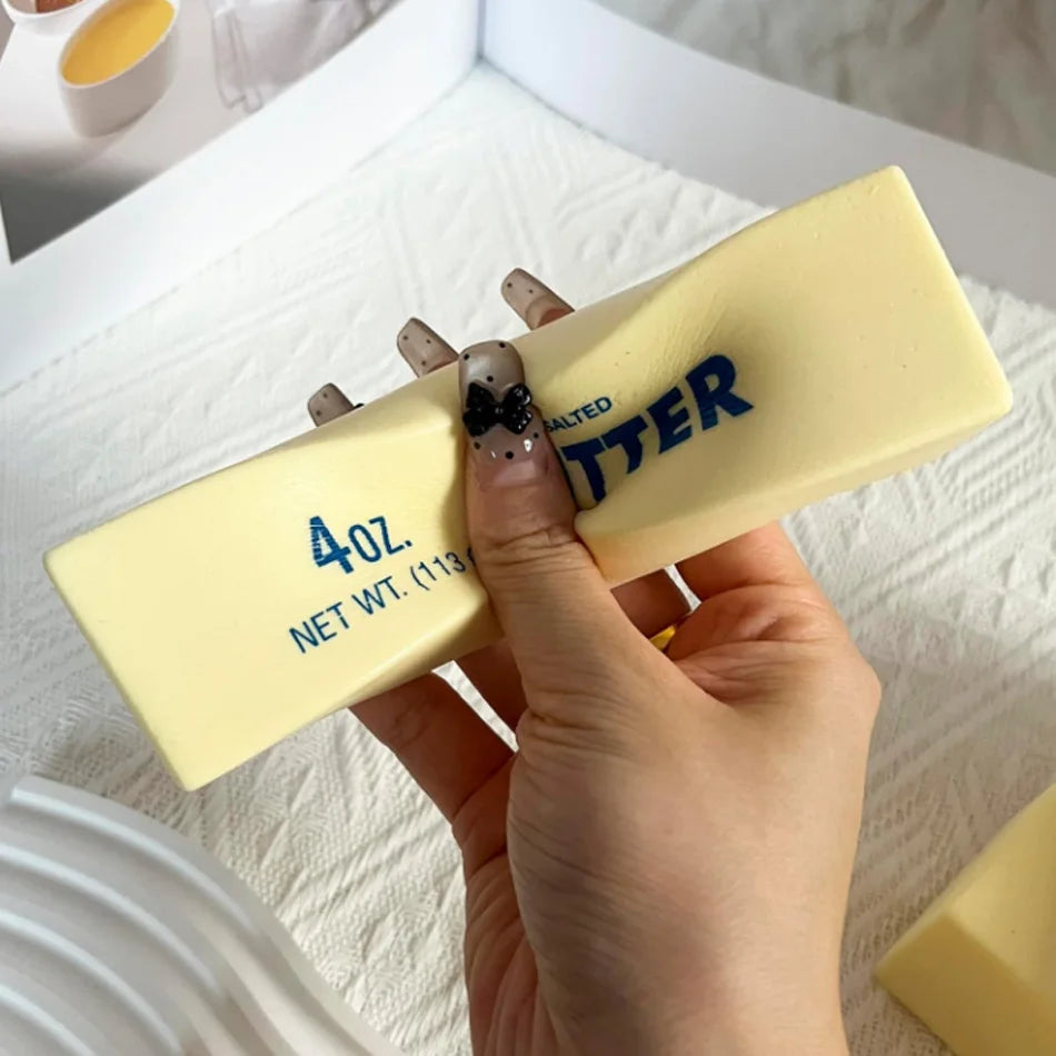 A hand holds a yellow butter squishy stress relief toy with a black charm on the finger.