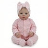A lifelike soft body baby doll with pink hair bow and pink outfit sitting on a white background.