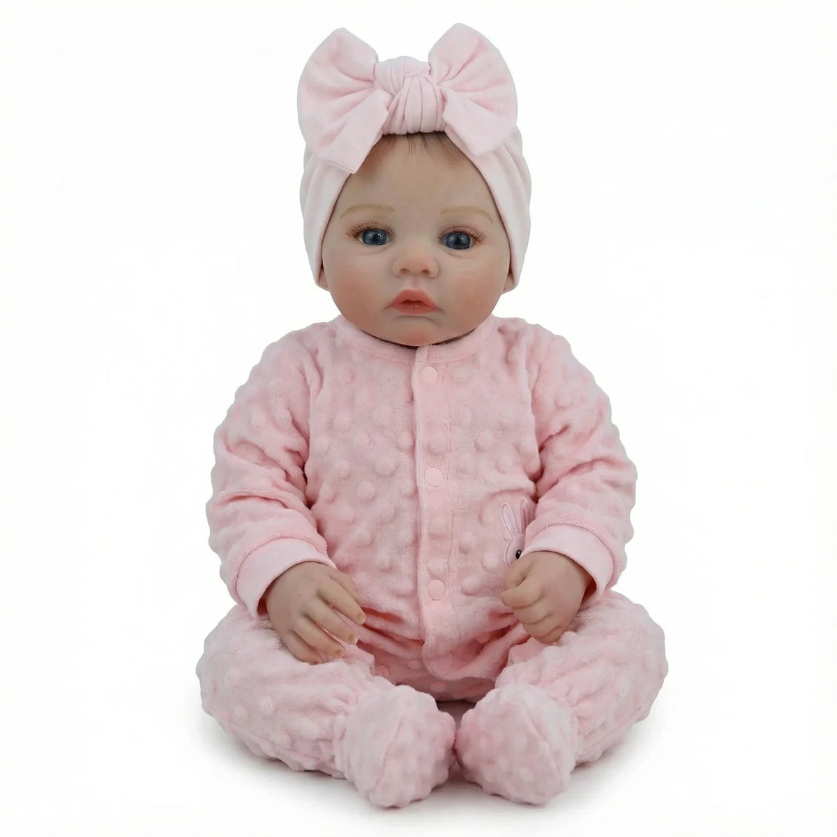 A lifelike soft body baby doll with pink hair bow and pink outfit sitting on a white background.