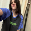 A person wearing a black t-shirt with green text and blue sleeves is shown indoors.